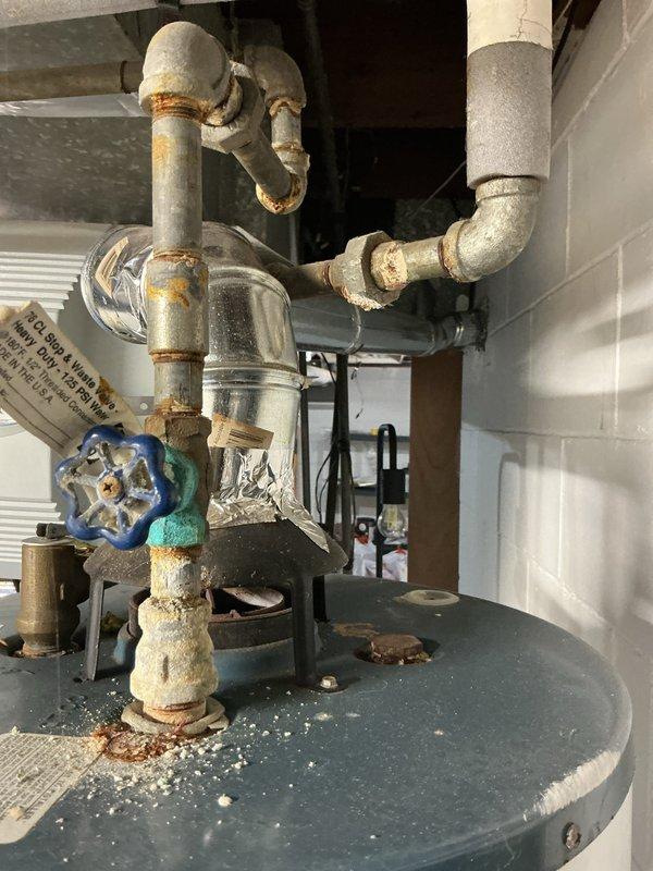 Responded to emergency water heater leak call. Upon inspection, found 10+ year old water heater with significant corrosion on inlet/outlet piping connections and fittings at top of tank. Blue-handled gate valve and surrounding threaded connections showed heavy rust and deterioration. Multiple pipe joints exhibited rust staining and mineral deposits, with insulation wrapping degraded and peeling on several sections. Pressure relief valve assembly inspected and found to be compromised due to age and corrosion. Tank top surface showed accumulated sediment and mineral deposits consistent with long-term use. Documented extensive corrosion damage to metal components and fittings throughout the water heater plumbing system, indicating need for replacement due to active leak and advanced deterioration of critical connections.