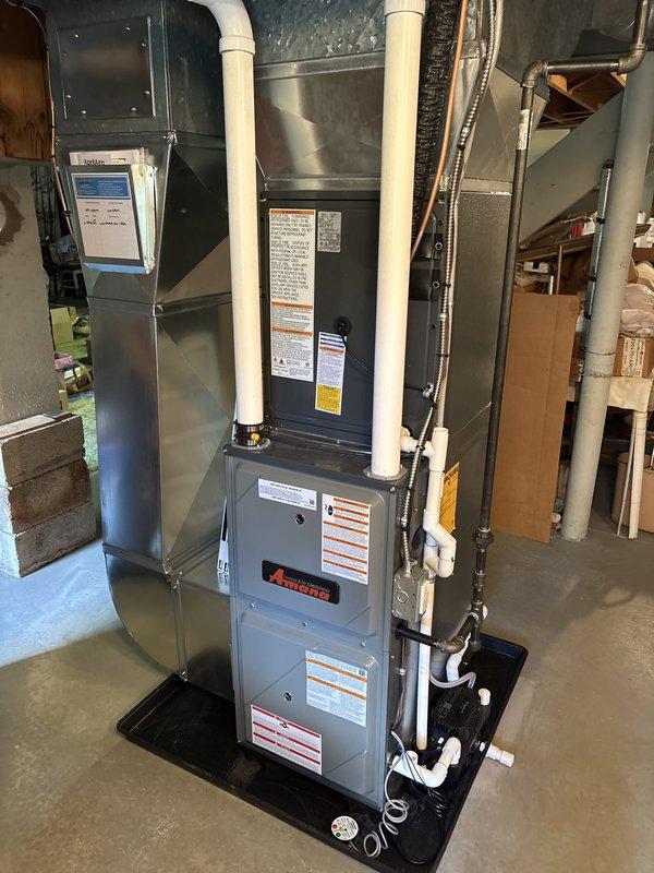 Completed full HVAC system installation including new furnace and air conditioning unit. Indoor furnace unit installed in basement/utility room with proper PVC intake and exhaust venting, mounted on drip pan with auxiliary condensate management system and connected ductwork. Outdoor condenser unit positioned on pad against exterior wall with electrical conduit and refrigerant lines properly routed through brick facade. All components tested and operational upon completion.