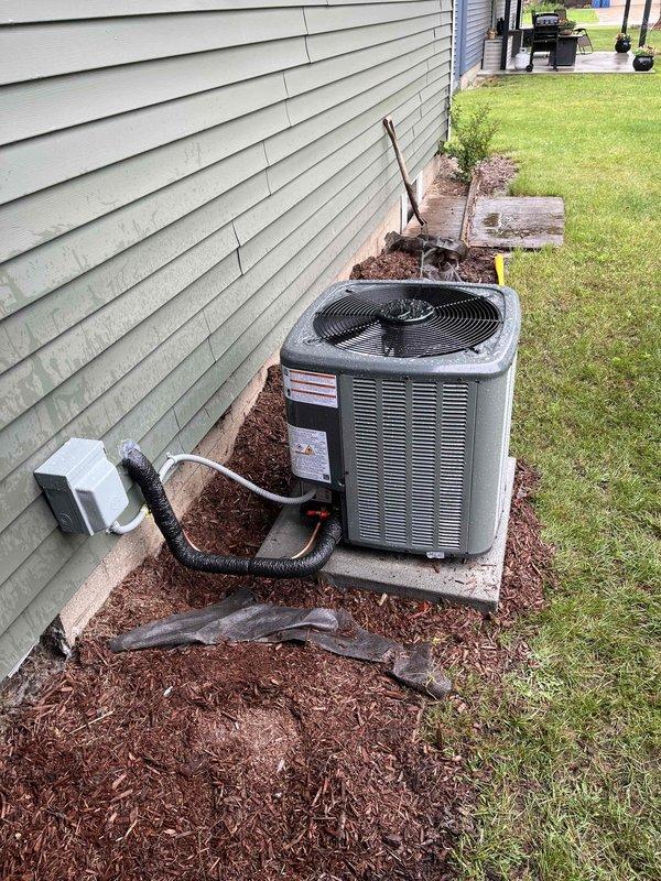 Completed full HVAC system installation including new furnace and air conditioning system. Installed grey metal furnace unit in basement on protective mat with proper venting using white PVC piping and copper refrigerant lines. Connected sheet metal ductwork plenum with reinforcement and integrated black flexible insulated ductwork to existing ceiling-mounted distribution system. Positioned outdoor condensing unit on concrete pad against exterior wall with proper clearance in mulched area. Installed electrical disconnect box on foundation near outdoor unit and ran black conduit for refrigerant line connection between indoor and outdoor components. All units properly labeled with safety warnings and specifications, system tested and verified operational.