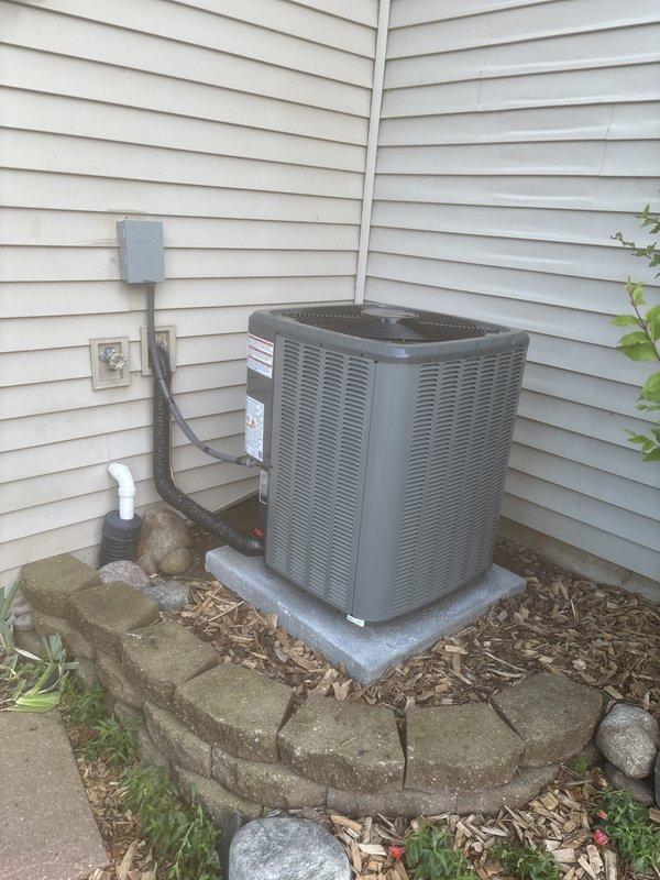 Completed full air conditioning system replacement at residential property. Removed existing outdoor condenser unit and installed new gray cylindrical AC condenser with horizontal ventilation slats and protective top grille. Unit positioned on existing concrete pad in corner location against beige vinyl siding. Connected electrical disconnect box to new unit via gray conduit, verified proper electrical connections and grounding. Installed white PVC condensate drain line with proper pitch and termination. Confirmed unit placement on elevated concrete pad with adequate clearance for airflow and service access. Landscaping area around unit maintained with mulch bed and decorative stone border featuring stacked gray pavers. Verified all manufacturer specifications, tested system operation, and confirmed proper cooling performance upon startup.
