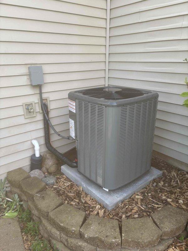 Completed full air conditioning system replacement at residential property. Removed existing outdoor condenser unit and installed new gray cylindrical AC condenser with horizontal ventilation slats and protective top grille. Unit positioned on existing concrete pad in corner location against beige vinyl siding. Connected electrical disconnect box to new unit via gray conduit, verified proper electrical connections and grounding. Installed white PVC condensate drain line with proper pitch and termination. Confirmed unit placement on elevated concrete pad with adequate clearance for airflow and service access. Landscaping area around unit maintained with mulch bed and decorative stone border featuring stacked gray pavers. Verified all manufacturer specifications, tested system operation, and confirmed proper cooling performance upon startup.