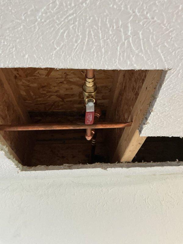 Cut access opening in ceiling to expose concealed plumbing. Located corroded copper hose bib with significant oxidation and patina buildup causing leak. Removed and replaced failed hose bib assembly with brass shut-off valve. Tested new installation for proper operation and verified no active leaks present.