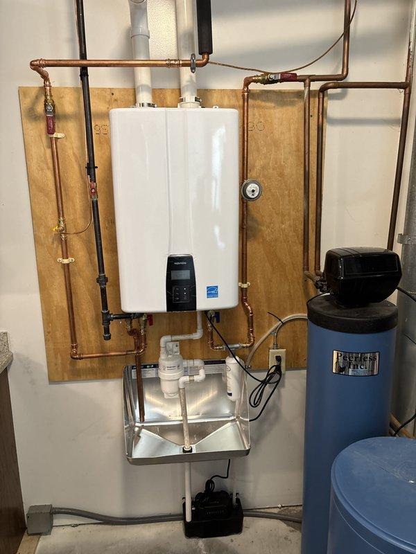 Completed full residential HVAC system replacement including installation of new furnace with vertical ductwork, air conditioning unit, and wall-mounted tankless water heater with condensate pan and PVC venting. Installed water treatment system consisting of two blue cylindrical filtration tanks alongside the tankless unit. All copper plumbing connections completed with pressure relief valves, gauges, and shutoff valves with red handles properly installed on water heater supply lines. Electrical connections established for all equipment including power adapter for tankless unit. System mounted on plywood backing board in mechanical room with proper venting, drainage, and safety components. All equipment tested and operational upon completion.