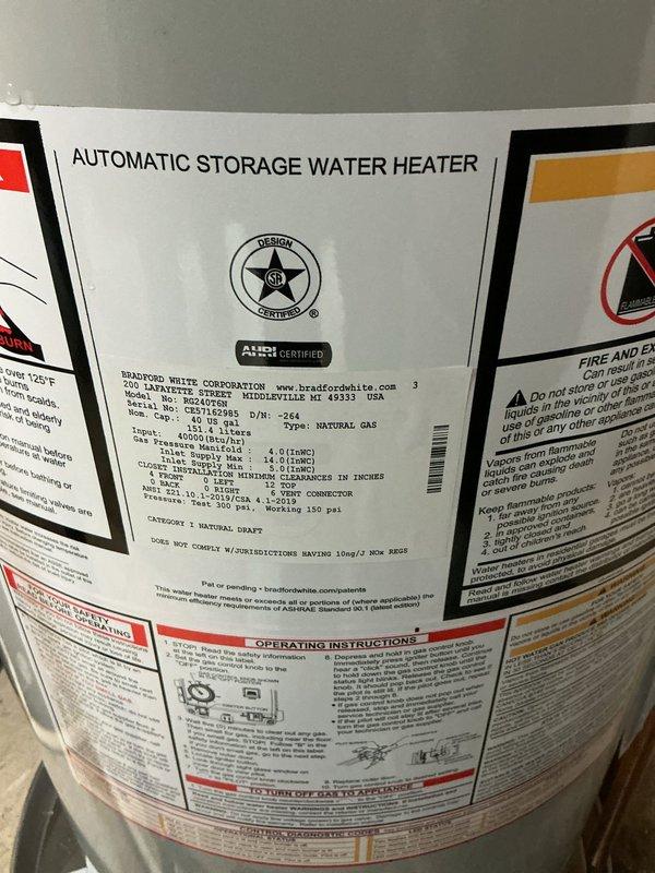 Completed installation of a 40-gallon tall natural gas Bradford White water heater rated at 40,000 BTU/HR in the mechanical room. Connected copper supply lines with brass fittings and white PVC venting to the top of the unit, ensuring proper integration with existing HVAC and plumbing systems. All manufacturer specifications, safety labels, and operating instructions are clearly visible and accessible on the installed unit.