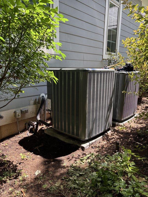 Completed full replacement of two outdoor air conditioning condensing units at residential property. Removed existing equipment and installed two new AC condensers side-by-side against exterior wall with gray horizontal siding. Units positioned on prepared ground surface with proper clearance for ventilation and serviceability. Installed electrical disconnect box on exterior wall for safe equipment shutdown capability. Ensured adequate airflow clearance around both units despite surrounding landscaping including nearby shrubs and vegetation. Verified proper pad placement, secure mounting, and appropriate spacing between units for optimal performance. All installations completed per manufacturer specifications and local code requirements.