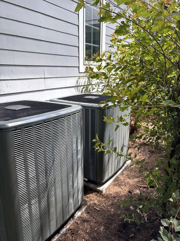Completed full replacement of two outdoor air conditioning condensing units at residential property. Removed existing equipment and installed two new AC condensers side-by-side against exterior wall with gray horizontal siding. Units positioned on prepared ground surface with proper clearance for ventilation and serviceability. Installed electrical disconnect box on exterior wall for safe equipment shutdown capability. Ensured adequate airflow clearance around both units despite surrounding landscaping including nearby shrubs and vegetation. Verified proper pad placement, secure mounting, and appropriate spacing between units for optimal performance. All installations completed per manufacturer specifications and local code requirements.