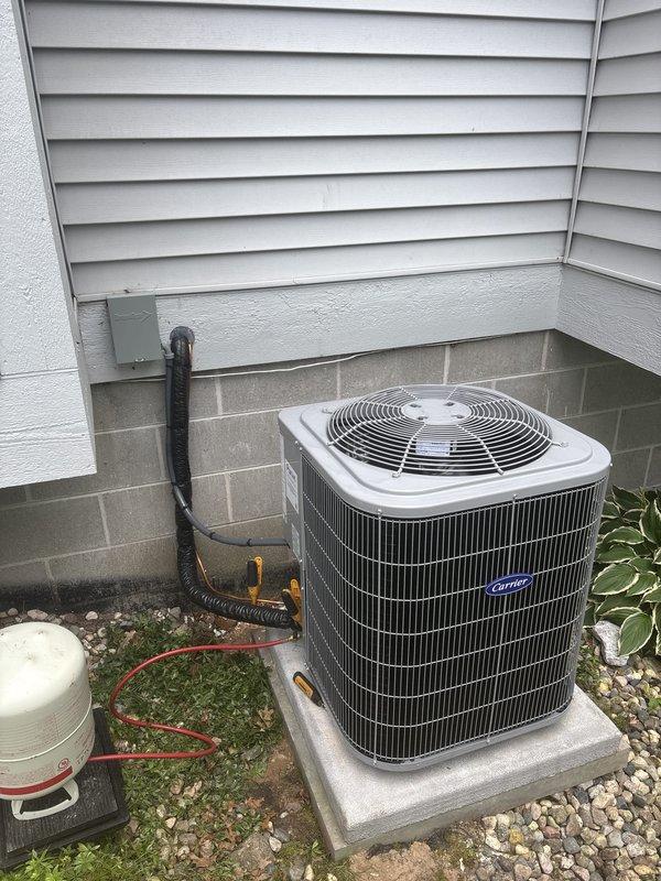 Completed full air conditioning system replacement at residential property. Removed existing outdoor condenser unit and installed new Carrier brand AC condenser on concrete pad against exterior wall with proper electrical connections via conduit mounted to siding. Unit positioned on decorative stone base for drainage and accessibility. Indoor components integrated with existing furnace and ductwork system in basement mechanical room, connecting to established humidifier and water heater infrastructure. All refrigerant lines, electrical connections, and condensate drainage properly installed and tested for optimal system performance.