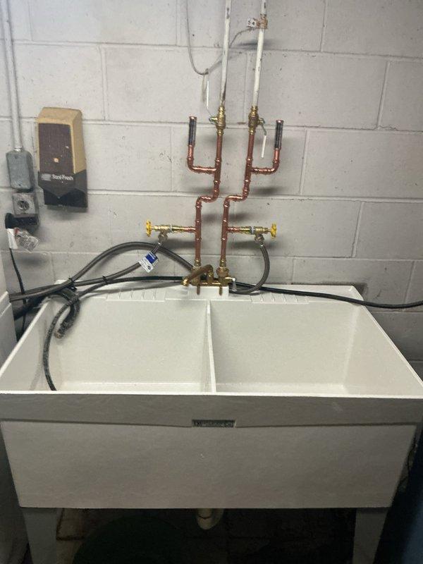 Completed installation of new white double-basin laundry sink with updated plumbing configuration including copper supply lines, brass boiler drain valves with yellow handles, PEX connections, flexible supply hoses, and new PVC P-trap drainage system mounted against concrete block wall in utility area.