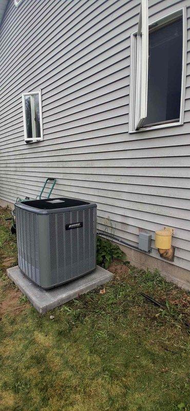 Completed full air conditioning system replacement at residential property. Removed existing condenser unit and installed new gray/silver AC condensing unit on existing concrete pad adjacent to house exterior with light tan vinyl siding. Connected refrigerant lines and electrical conduit from new condenser to indoor air handler. Verified proper placement on level concrete base, tested all electrical connections, and confirmed system operation. Unit installation includes protective mesh grille housing and manufacturer specifications compliance. All refrigerant lines properly insulated and secured to building structure.