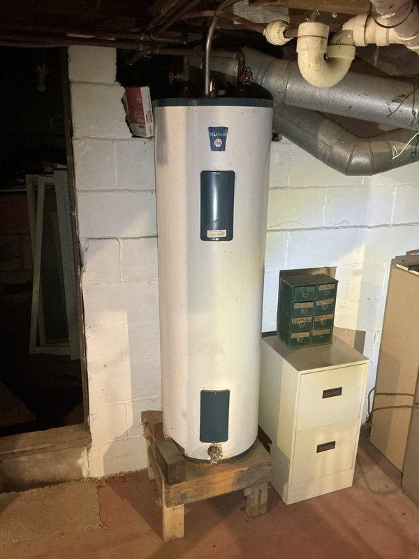 Conducted operational assessment of residential HVAC system and water heater in basement utility area at 3PM. Documented existing beige furnace unit with flexible metallic ductwork connections and large white cylindrical water heater mounted on wooden platform base. Photographed current equipment configuration, ductwork routing, and mechanical installations to provide comprehensive estimate for air conditioning system evaluation and potential service requirements.
