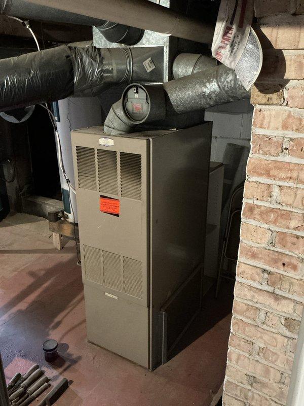 Conducted operational assessment of residential HVAC system and water heater in basement utility area at 3PM. Documented existing beige furnace unit with flexible metallic ductwork connections and large white cylindrical water heater mounted on wooden platform base. Photographed current equipment configuration, ductwork routing, and mechanical installations to provide comprehensive estimate for air conditioning system evaluation and potential service requirements.