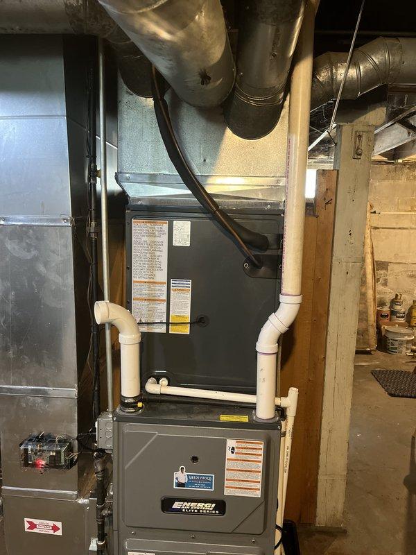 Completed full HVAC system replacement including outdoor condensing unit and indoor air handler. Removed existing equipment and installed new gray condensing unit on concrete pad adjacent to residential structure with proper clearances maintained from deck and building exterior. Installed new air handler in basement mechanical room with integration to existing furnace system. Ran new condensate drainage lines using white PVC piping with proper pitch and termination. Connected refrigerant lines between outdoor and indoor units with proper insulation and routing. Verified all electrical connections, tested system startup and operation, and confirmed proper airflow and temperature differential across evaporator coil.
