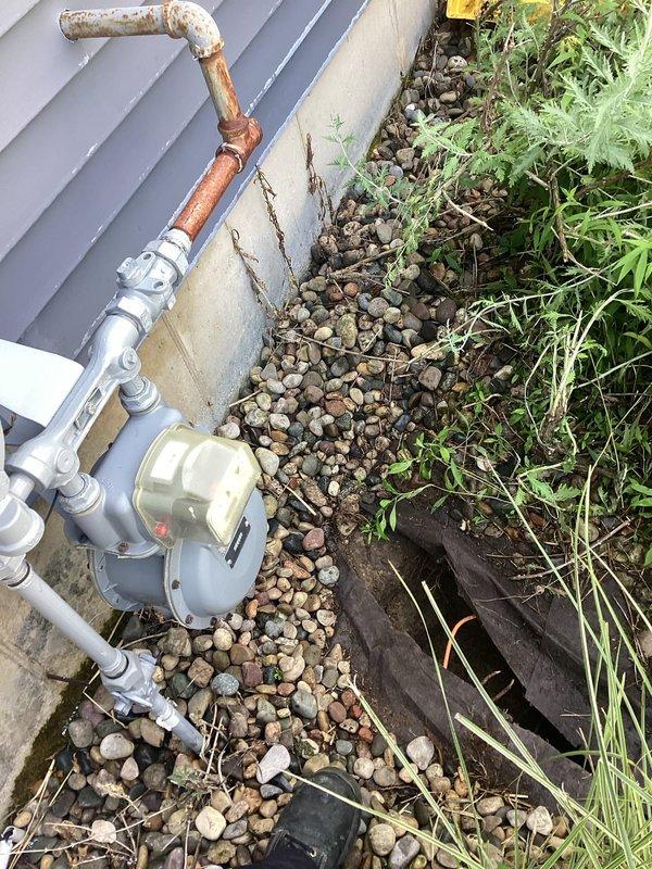 Conducted on-site evaluation for gas line installation to service pool heater. Assessed existing gas meter assembly and piping infrastructure located at building exterior, noting copper and galvanized metal pipe configuration with visible weathering and corrosion on existing components. Evaluated routing requirements from current gas meter location through yard area to proposed pool heater installation point.

Documented existing utility connections and meter placement against building foundation. Identified excavation pathway through landscaped area between evergreen shrubs where new gas line would need to be installed. Photographed trench route and ground conditions to determine appropriate pipe sizing, depth requirements, and material specifications for compliant installation. Assessed soil conditions and existing landscape features that would impact trenching work and restoration requirements upon project completion.