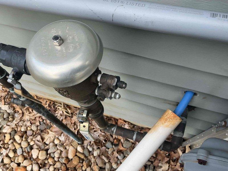 Conducted plumbing project estimate for property with dual water treatment systems—existing water softener and Gordon water system. Evaluated feasibility of removing one system to optimize space and assessed requirements for installing new laundry room sink. Documented exterior plumbing infrastructure including corroded copper piping with temporary patch repairs, exterior-mounted pressure regulation equipment, and ground-level utility connections with PVC drainage lines. Photographed existing conditions to provide comprehensive assessment for system consolidation and laundry room addition.