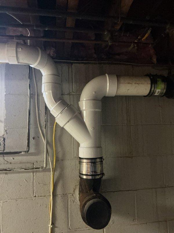 Completed repipe installation in basement area, replacing deteriorated metal piping with new white PVC drainage system. Installed proper fittings including 90-degree elbows and Y-junction connections, transitioning from new PVC to existing cast iron waste line using rubber coupling adapter. Work performed beneath floor joists with proper routing and support to ensure code-compliant installation and optimal drainage flow.