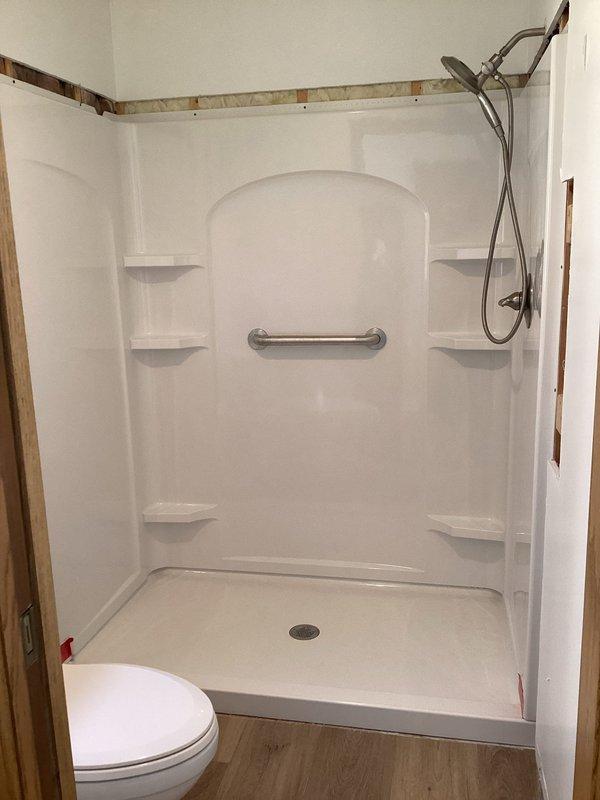 Completed installation of walk-in shower unit with white fiberglass surround panels, including integrated corner shelving on both sides and centered arched alcove in back wall. Installed chrome shower fixture system with handheld showerhead attachment and flexible hose, mounted stainless steel grab bar within arched recess for safety accessibility, and ensured proper drainage with centered floor drain in shower base.
