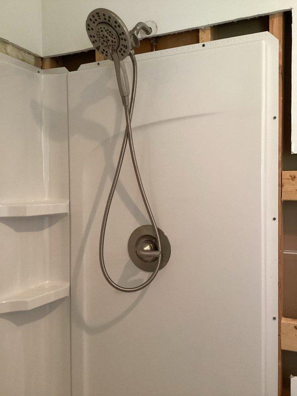 Completed installation of walk-in shower unit with white fiberglass surround panels, including integrated corner shelving on both sides and centered arched alcove in back wall. Installed chrome shower fixture system with handheld showerhead attachment and flexible hose, mounted stainless steel grab bar within arched recess for safety accessibility, and ensured proper drainage with centered floor drain in shower base.