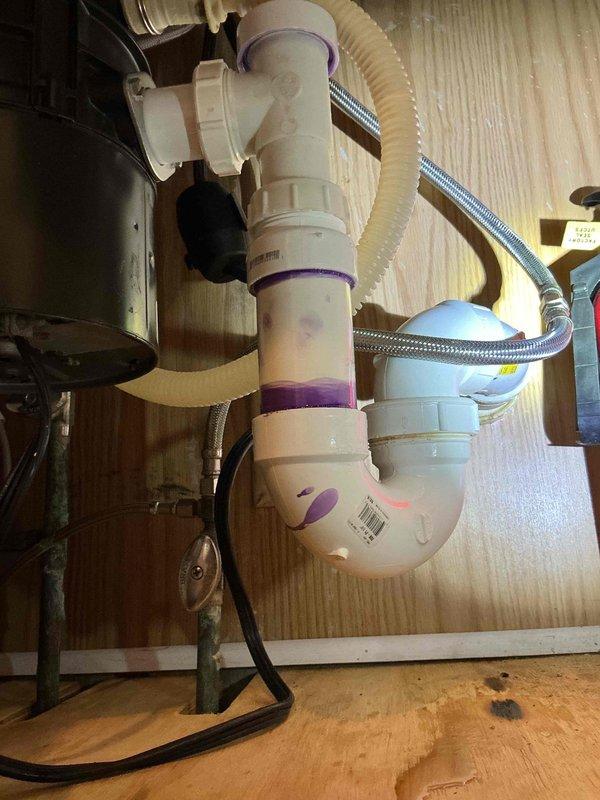 Responded to a kitchen sink leak that had been present for approximately two months in a residential property constructed in 1980. Upon inspection, identified the source of the leak at the under-sink P-trap assembly with purple rubber couplings connecting white PVC drain components. Examined the complete drainage configuration including connections to the garbage disposal unit via corrugated flexible hose and verified the integrity of braided stainless steel water supply lines. Tightened all coupling connections at the P-trap joints and inspected for proper seal integrity. Tested the system by running water through the sink and disposal to confirm leak elimination. Verified all hose clamps were properly secured on both the drain hose and supply lines. Cleaned the cabinet interior and removed moisture residue from previous leaking. System now functioning properly with no active leaks detected.