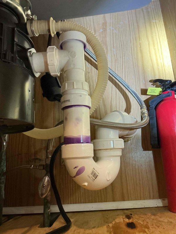 Responded to a kitchen sink leak that had been present for approximately two months in a residential property constructed in 1980. Upon inspection, identified the source of the leak at the under-sink P-trap assembly with purple rubber couplings connecting white PVC drain components. Examined the complete drainage configuration including connections to the garbage disposal unit via corrugated flexible hose and verified the integrity of braided stainless steel water supply lines. Tightened all coupling connections at the P-trap joints and inspected for proper seal integrity. Tested the system by running water through the sink and disposal to confirm leak elimination. Verified all hose clamps were properly secured on both the drain hose and supply lines. Cleaned the cabinet interior and removed moisture residue from previous leaking. System now functioning properly with no active leaks detected.
