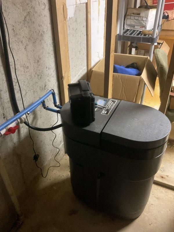 Responded to service call for non-functioning water softener system. Upon inspection, evaluated the brine tank and control unit setup in the basement utility area. Assessed the system components including the resin tank, control valve assembly, and brine tank configuration to diagnose operational issues. Documented existing installation conditions with photographic evidence showing the water softener equipment and surrounding mechanical space for service records.