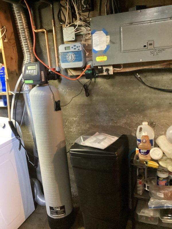 Removed existing water treatment equipment from basement utility area and completed installation of new Sterling high-efficiency water softener system with integrated whole house sediment filter. Configured copper supply piping with brass compression fittings and mounted white sediment filter housing to concrete wall with proper inlet/outlet connections. System installed, tested, and left operational with all components properly positioned and secured.