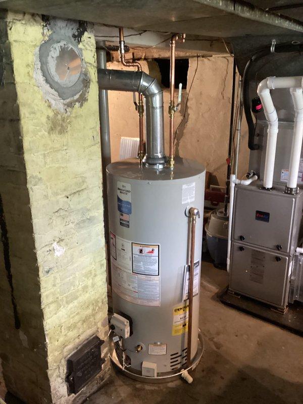 Responded to emergency call for leaking water heater in residential basement. Upon inspection, found 15-year-old 40-gallon water heater actively leaking and requiring immediate replacement. Unit had reached end of serviceable life with visible deterioration. Documented existing installation configuration including copper supply lines, PVC venting system, and proximity to furnace unit in mechanical room. Photographed current setup showing water heater connections, surrounding plumbing infrastructure, and basement utility area layout to facilitate replacement planning. Provided estimate for complete water heater replacement to restore hot water service.