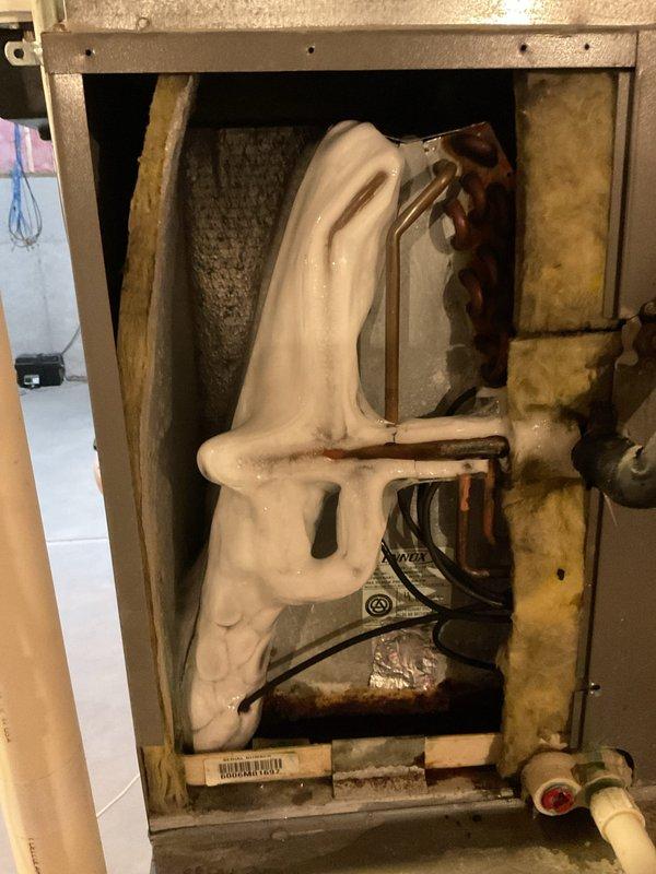 # Cooling System Airflow Restriction - Evaporator Coil Icing

Responded to service call for residential cooling system unable to maintain temperature below 80°F. Upon inspection of 19-year-old Lennox unit, discovered severe ice accumulation on evaporator coil assembly in air handler. The coil exhibited heavy frost buildup covering the refrigerant lines in a serpentine pattern, indicating restricted airflow or possible refrigerant charge issues. Documented equipment condition with photographs showing the frozen coil through the access panel and the basement installation location with visible moisture on floor near unit. The ice formation pattern and system age suggest potential causes including dirty air filter, blocked return air, low refrigerant charge, or failing blower motor. Recommended system evaluation after coil defrost to determine root cause of airflow restriction and prevent recurrence of freezing condition.