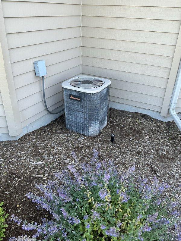 Responded to service call for non-operational air conditioning system at residential property. Upon arrival, conducted comprehensive evaluation of existing HVAC equipment including exterior Amana condenser unit with protective grille housing and interior Goodman furnace system with associated ductwork and Honeywell humidifier. Performed diagnostic assessment of both outdoor condensing unit and indoor air handler components to determine cause of system failure. Documented current equipment condition, configuration, and installation setup through photographic records showing condenser placement on mulched pad with electrical disconnect box, as well as mechanical room layout featuring furnace, plenum assembly, galvanized ductwork, and PVC drainage piping. Prepared detailed estimate for necessary repairs or equipment replacement options to restore cooling functionality to the residence.