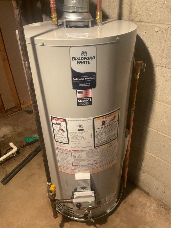 Successfully replaced and installed a 40-gallon natural gas Bradford White water heater in basement utility area. The new short-profile unit was properly positioned against the concrete block wall with all necessary plumbing connections completed, including galvanized and copper pipe fittings at the top of the unit. Gas line connection was established with safety valve installed and verified for proper operation.