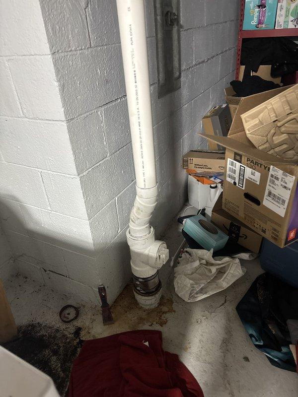 Responded to blocked drain call in basement utility area. Upon inspection, found drainage system with PVC piping running along corner walls and floor level connections. Assessed plumbing configuration including U-trap assembly with black pipes emerging from floor. Cleared blockage in drain line, restoring proper flow and drainage function. Verified system operation and confirmed all pipes were draining correctly upon completion.