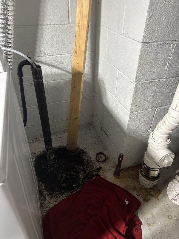 Responded to blocked drain call in basement utility area. Upon inspection, found drainage system with PVC piping running along corner walls and floor level connections. Assessed plumbing configuration including U-trap assembly with black pipes emerging from floor. Cleared blockage in drain line, restoring proper flow and drainage function. Verified system operation and confirmed all pipes were draining correctly upon completion.