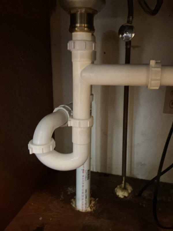 Replaced kitchen drain line assembly beneath sink, including P-trap configuration and vertical drain pipe that showed evidence of water damage and deterioration at floor penetration. Installed new white PVC drainage system with proper trap configuration, chrome tailpiece connection to sink drain, and extended drainage piping through floor structure to connect with existing waste lines in basement/crawlspace below. All connections sealed and tested for proper drainage flow.