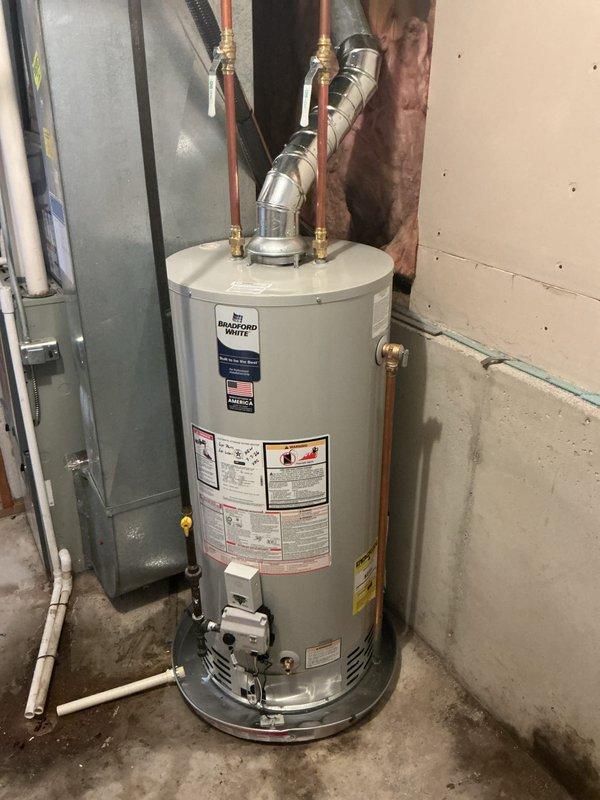 Completed installation of 40-gallon natural gas Bradford White water heater with 6-inch vent diameter and short configuration. Connected copper supply lines with brass fittings, installed flexible water connections, configured gas valve assembly and controls, and installed metallic vent pipe with proper 90-degree elbow termination through ceiling penetration. Unit installed in basement utility area on concrete floor with all safety labels visible and accessible, meeting code requirements for residential water heating systems.