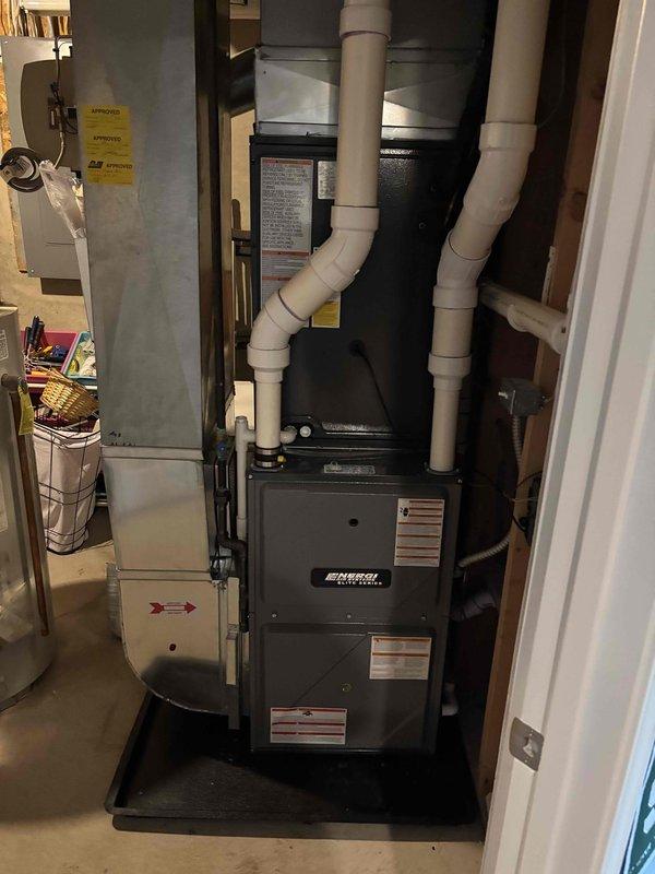 Completed full HVAC system replacement including outdoor condensing unit and indoor air handler with ENERGY STAR rated furnace. Removed existing equipment and installed new gray-green condensing unit on concrete pad with proper clearances from structure. Mounted electrical disconnect box on exterior siding and ran conduit from house to unit per code requirements. Inside installation included dark cabinet furnace with integrated humidifier system, white PVC exhaust and intake venting configured in dual-pipe arrangement, and condensate drain pan positioned beneath unit for proper water management. Verified all electrical connections, refrigerant line sets, and drainage systems operational. System tested and confirmed proper operation of heating and cooling cycles.