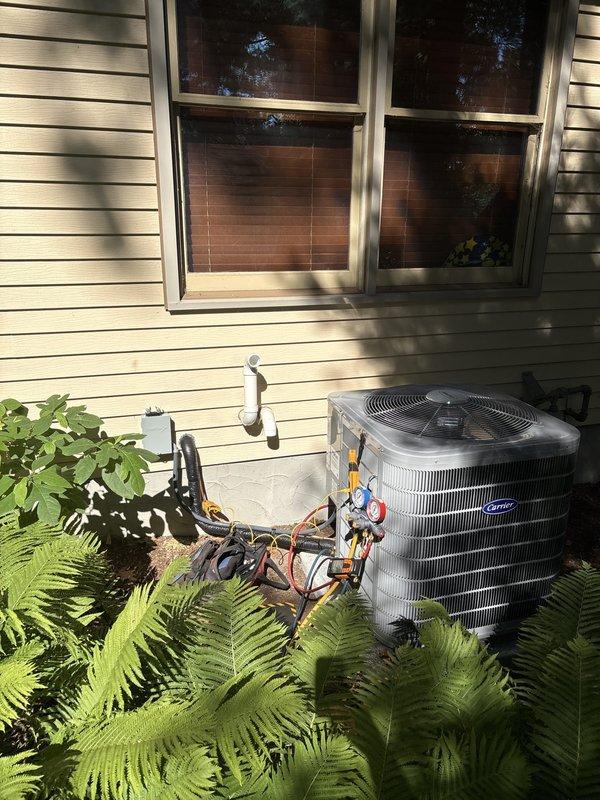 Successfully completed full air conditioning system replacement at residential property. Removed existing outdoor condensing unit and installed new Carrier brand condenser with proper refrigerant line connections and condensate drainage routing. Unit positioned on stable pad with copper refrigerant lines and white PVC condensate piping properly secured to exterior wall. Interior basement installation included integration with existing furnace system, ensuring proper plenum connections and ductwork configuration. All refrigerant lines insulated appropriately, condensate drainage system installed with PVC piping to floor drain, and electrical connections completed per manufacturer specifications. System tested for proper operation, refrigerant charge verified, and airflow confirmed throughout ductwork. Installation site left clean with landscaping undisturbed around exterior unit.