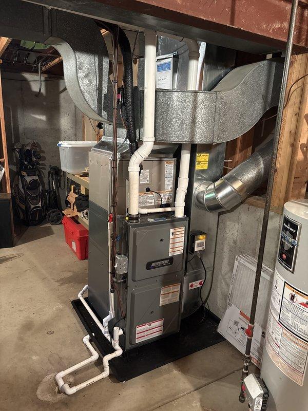Successfully completed full air conditioning system replacement at residential property. Removed existing outdoor condensing unit and installed new Carrier brand condenser with proper refrigerant line connections and condensate drainage routing. Unit positioned on stable pad with copper refrigerant lines and white PVC condensate piping properly secured to exterior wall. Interior basement installation included integration with existing furnace system, ensuring proper plenum connections and ductwork configuration. All refrigerant lines insulated appropriately, condensate drainage system installed with PVC piping to floor drain, and electrical connections completed per manufacturer specifications. System tested for proper operation, refrigerant charge verified, and airflow confirmed throughout ductwork. Installation site left clean with landscaping undisturbed around exterior unit.