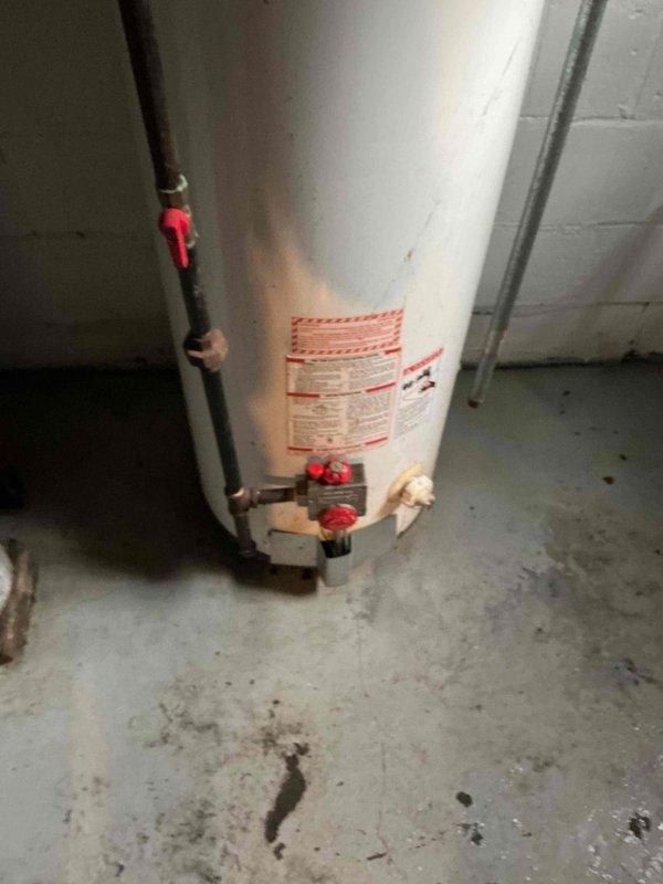 Responded to service call for 5-year-old water heater leaking from bottom. Upon inspection, identified active leak at base of unit near plumbing connections and valve assembly. Documented existing conditions including staining on concrete floor around heater base and configuration of inlet/outlet piping with red ball valves and copper fittings. Photographed water heater installation, mechanical room environment, and associated HVAC ductwork for service records.