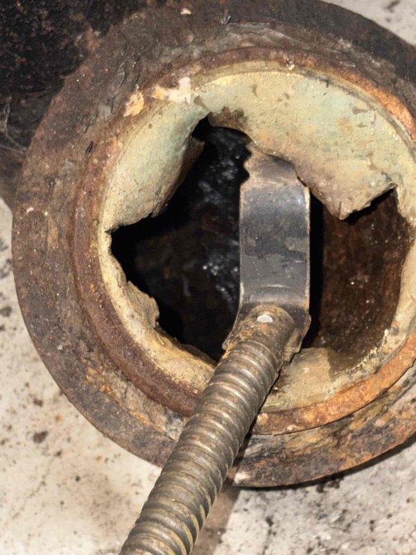 Responded to emergency main floor drain backup causing complete loss of water usage throughout residence due to basement flooding. Upon arrival, located floor drain in basement utility area adjacent to laundry appliances. Inspection revealed severely corroded drain pipe opening with extensive rust deterioration and compromised pipe integrity. The circular drain opening showed significant metal oxidation with exposed deteriorated insulation material around the interior rim. Flexible metal conduit connection displayed signs of age and moisture exposure. Documented existing conditions showing concrete floor discoloration, dirt accumulation around drain area, and corroded metal fittings at pipe base. The 1960s-era stick-built home's original plumbing infrastructure exhibited advanced corrosion consistent with decades of use and moisture exposure. Photographed drain access point and surrounding utility space for record purposes. Assessed drainage system to determine source of backup and flo