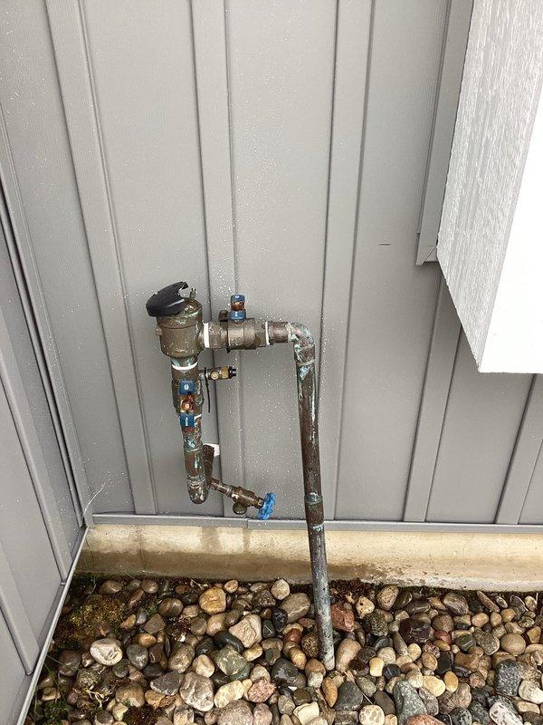 Responded to plumbing leak in backflow prevention system mounted on exterior wall. Upon inspection, identified active leak at brass backflow preventer assembly with multiple ball valves and copper piping connections. Assessed the outdoor plumbing installation which showed signs of weathering and exposure to elements. Repaired leak by tightening compromised fittings and replacing worn components as needed. Tested all blue-handled ball valves for proper operation and verified system integrity. Confirmed backflow preventer functioning correctly with no active leaks upon completion. System restored to full operational status.