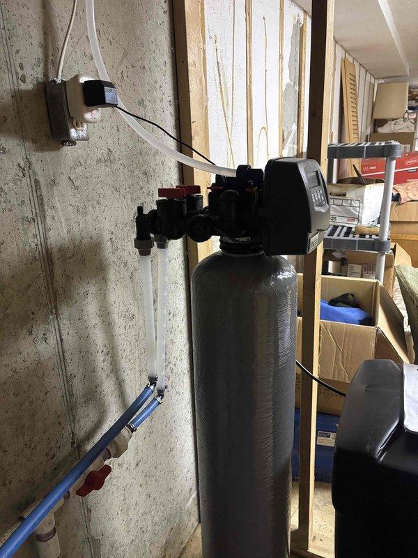Completed installation of Sterling Water Treatment water softening system in utility area. Mounted control head with digital display panel on gray cylindrical softener tank, connected power supply to wall outlet, and routed blue drain line for proper discharge. System programmed and tested, confirmed operational status with control panel displaying time and settings. Installation completed with all plumbing connections secure and unit ready for service.