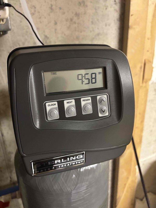 Completed installation of Sterling Water Treatment water softening system in utility area. Mounted control head with digital display panel on gray cylindrical softener tank, connected power supply to wall outlet, and routed blue drain line for proper discharge. System programmed and tested, confirmed operational status with control panel displaying time and settings. Installation completed with all plumbing connections secure and unit ready for service.