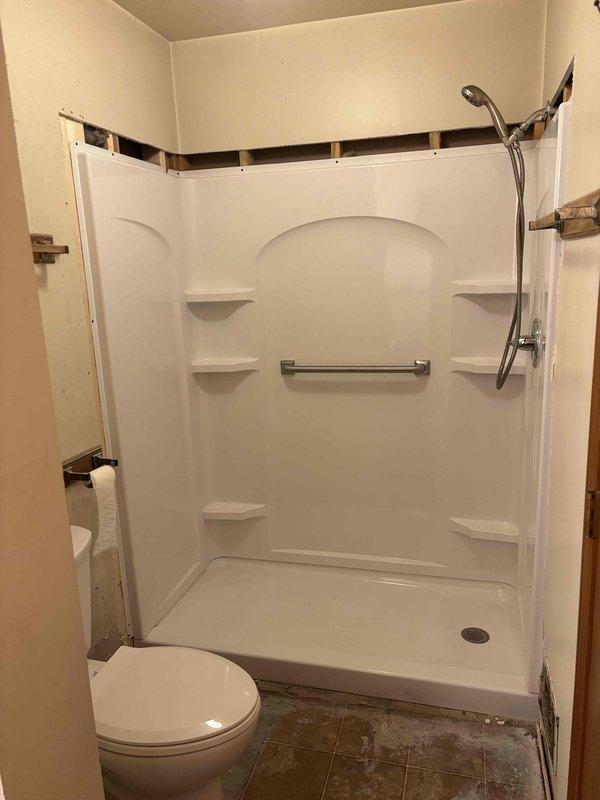 Completed installation of prefabricated white shower unit with integrated shelving, chrome grab bar, and flexible shower head assembly, including proper drain connection and threshold sealing. Installed new two-piece toilet with tank and bowl configuration, ensuring proper wax ring seal and water supply connection. Work performed in bathroom undergoing renovation with coordination around ongoing wall finishing and construction activities.