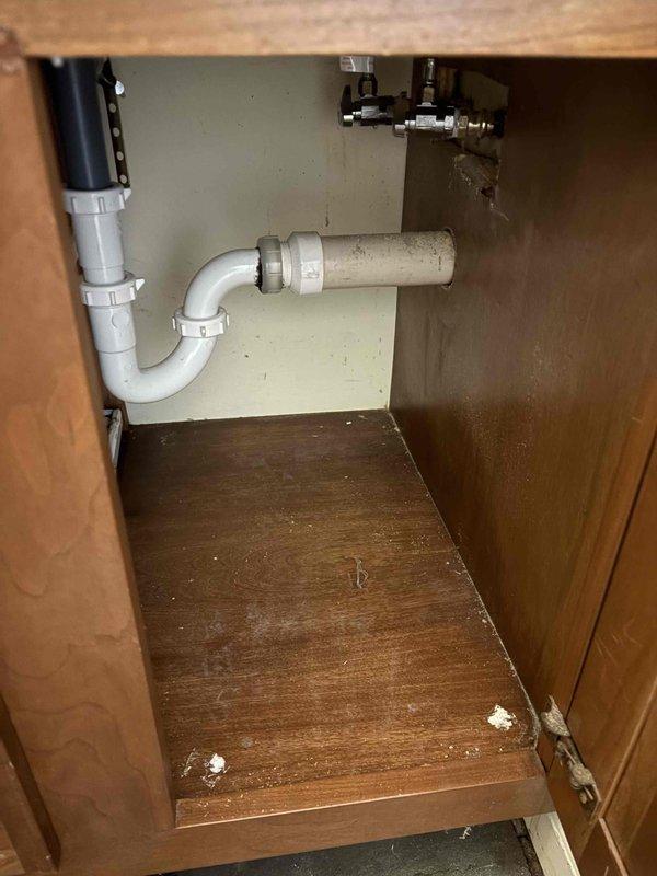 Responded to plumbing leak in basement of 1970s-era residential property where water was discovered on floor in rarely used room. Upon inspection, traced water source to leaking pipe connections in exposed basement plumbing system. Examined PVC drainage piping secured to wooden framing with metal strapping brackets and inspected under-sink cabinet plumbing including P-trap assembly, shut-off valves, and supply line connections. Documented conditions with photographs showing exposed framing, ductwork, and plumbing infrastructure in crawl space area, as well as cabinet interior with visible wear and moisture indicators. Confirmed leak location and provided assessment of aging plumbing system requiring repair to prevent further water damage.