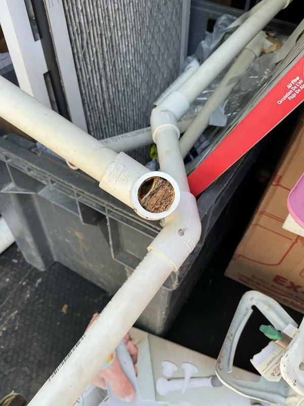 Responded to water leak complaint below air conditioning unit in basement. Upon inspection, discovered significant sediment and debris accumulation blocking the condensate drain line, causing water to overflow from the system. Documented condition of PVC drainage piping showing brown discoloration and buildup inside the drain line. Air handler unit confirmed operational and cooling properly despite drainage issue. Photographed affected components including T-junction connection and interior pipe condition to document extent of blockage requiring clearing to restore proper condensate drainage function.
