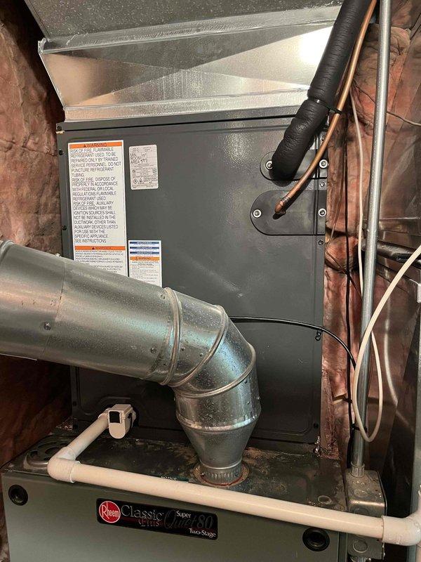 Complete residential HVAC system replacement performed, including outdoor condensing unit and indoor air handler installation. Removed existing equipment and installed new olive green outdoor unit on concrete pad against exterior brick wall. Mounted unit includes proper electrical disconnect box with black flexible conduit running vertically along wall for code-compliant power supply. Unit positioned in side yard utility area with appropriate clearances maintained.

Indoor air handler installed in attic space above existing Rheem Classic Plus Series 80 Two-Stage furnace. Connected refrigerant lines properly insulated and secured with mounting brackets. Integrated new air handler with existing galvanized ductwork system using proper elbow connections. All refrigerant lines charged and tested for proper operation. System startup completed with verification of cooling cycle, airflow, and thermostat communication. Installation meets manufacturer specifications and local building codes.