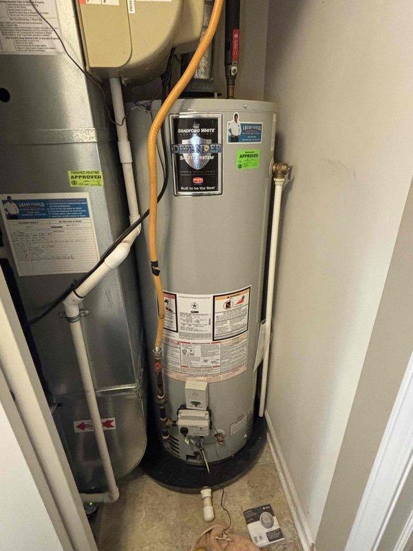 Responded to report of water collecting in drain pan under 5-year-old Bradford White water heater. Upon inspection, verified water heater installation and connections in residential utility closet. Documented equipment condition with photos showing Bradford White Defender Safety System model with standard plumbing connections, gas line, and HVAC equipment in confined space. All visible components including water lines, gas connections, and drain pan were assessed for proper operation and potential leak sources.