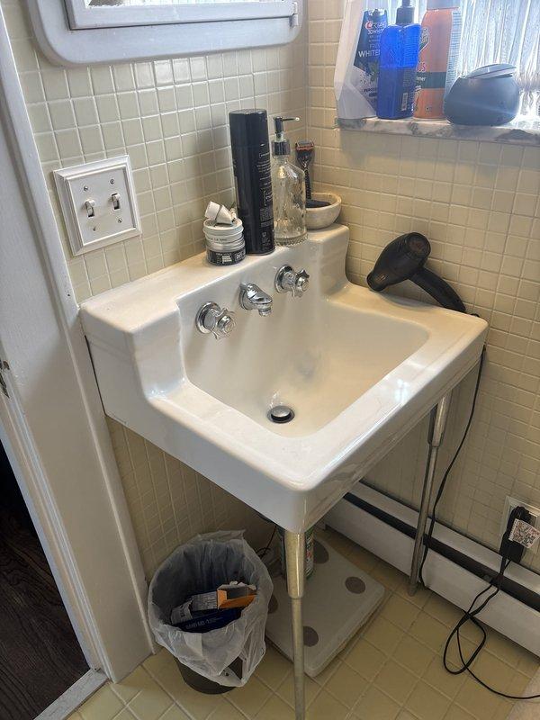 Conducted comprehensive site assessment for plumbing renovation project in century-old residential property. Evaluated existing bathroom infrastructure including wall-mounted pedestal sink with chrome fixtures, exposed electrical wiring along baseboards, and cream-colored tile work requiring updates. Documented condition of bathroom fixtures and photographed two new white vanity cabinets with recessed panel doors and drawer storage awaiting installation. Provided detailed project estimate for complete pipe replacement and bathroom fixture installation to modernize plumbing system while addressing age-related infrastructure concerns in historic home.