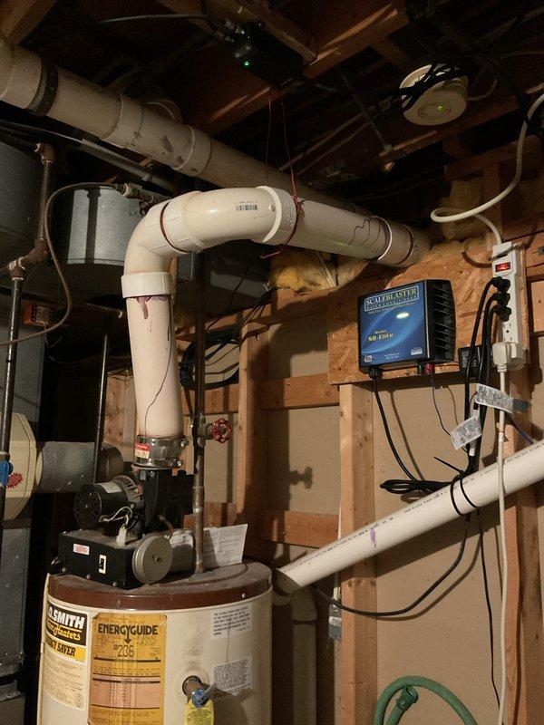 Provided estimate for water heater replacement in unfinished basement utility area. Assessed existing white tank-style water heater with current plumbing configuration including white PVC venting system, copper supply lines, and recirculation pump with electronic controls. Documented existing installation featuring ScaleBlaster water conditioning system, gray wall-mounted HVAC or water treatment unit, and black circulation pump at base of current heater. Photographed complete plumbing layout including complex PVC piping arrangements with multiple elbows and joints, aged copper piping with visible patina, and blue electronic controller for pump system. Reviewed accessibility within exposed wooden framing and evaluated requirements for new water heater installation to match existing connections and maintain proper venting through current PVC system.