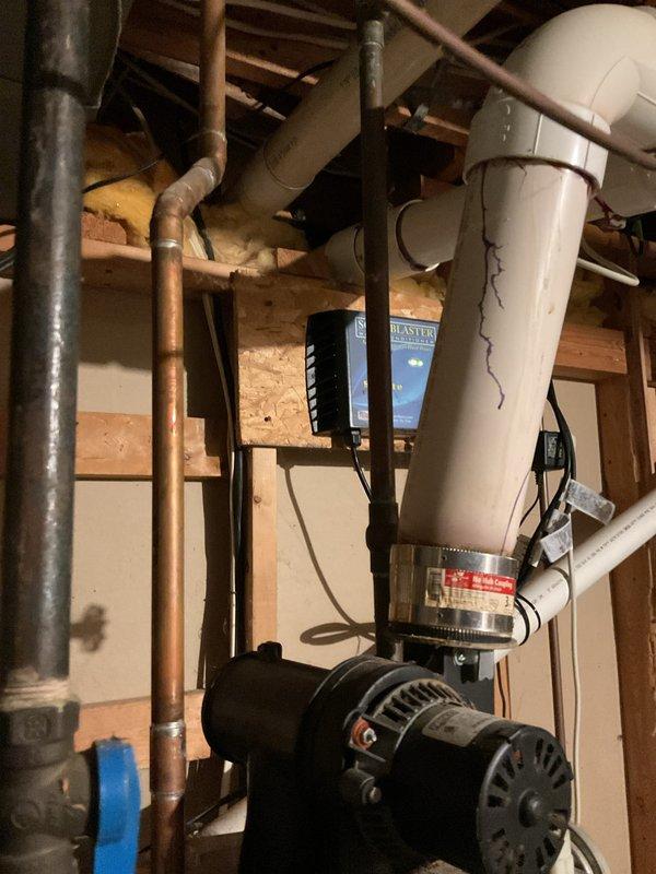 Provided estimate for water heater replacement in unfinished basement utility area. Assessed existing white tank-style water heater with current plumbing configuration including white PVC venting system, copper supply lines, and recirculation pump with electronic controls. Documented existing installation featuring ScaleBlaster water conditioning system, gray wall-mounted HVAC or water treatment unit, and black circulation pump at base of current heater. Photographed complete plumbing layout including complex PVC piping arrangements with multiple elbows and joints, aged copper piping with visible patina, and blue electronic controller for pump system. Reviewed accessibility within exposed wooden framing and evaluated requirements for new water heater installation to match existing connections and maintain proper venting through current PVC system.