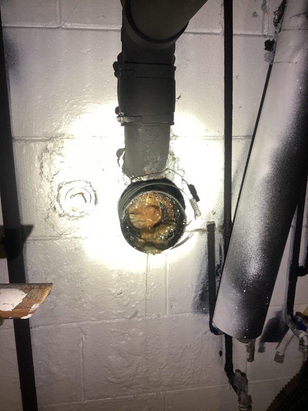 Responded to drain problem at property. Upon inspection, found corroded metal drain flange mounted to basement wall with significant rust buildup and mineral deposits obstructing the interior opening. Assessed plumbing assembly including vertical drain pipe connection showing signs of age, water damage, and debris accumulation around connection point. Documented existing conditions of wall-mounted drainage system and surrounding utility infrastructure for service records.